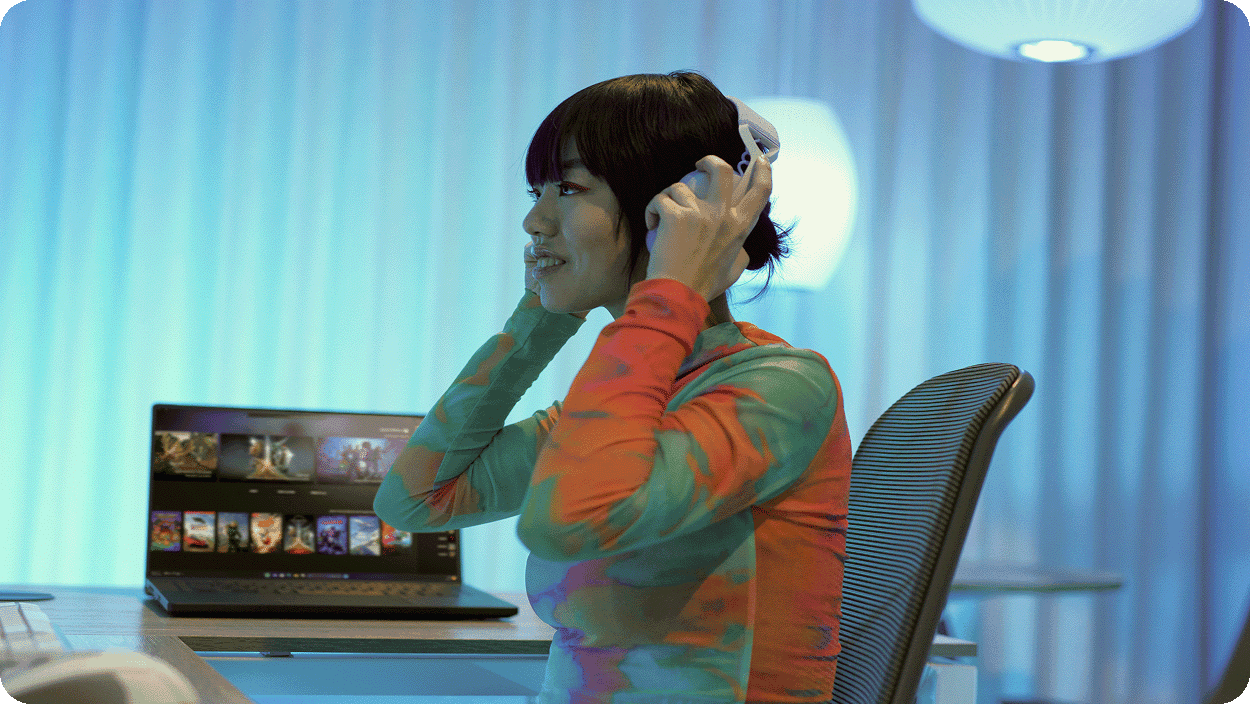 A woman sits at a desk in a softly lit room with blue tones, placing headphones on her head while facing a laptop displaying multiple images or videos.